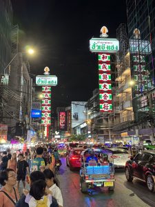 Tuk-tuk and evening traffic beneath tall red-and-green illuminated signs on Yaowarat Road in Bangkok’s Chinatown.