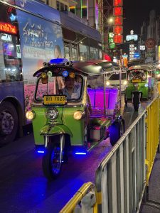Green tuk-tuk with blue and purple lights beside a bus on Yaowarat Road in Bangkok’s Chinatown at night.